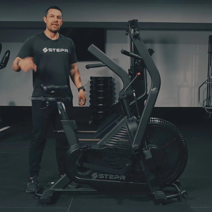 Man standing in a gym with exercise equipment, wearing a branded shirt.
